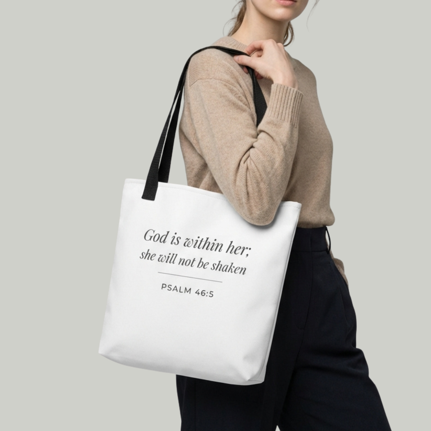 Lifestyle photo of a model holding a white tote bag featuring ‘God is within her; she will not be shaken’ (Psalm 46:5) in dark gray.