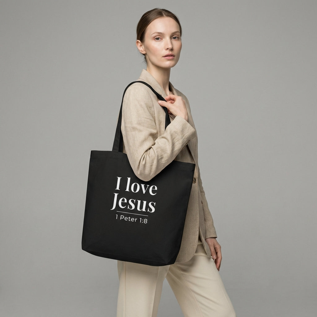 Model holding a black cotton tote bag printed with “I love Jesus” and “1 Peter 1:8” in white text.