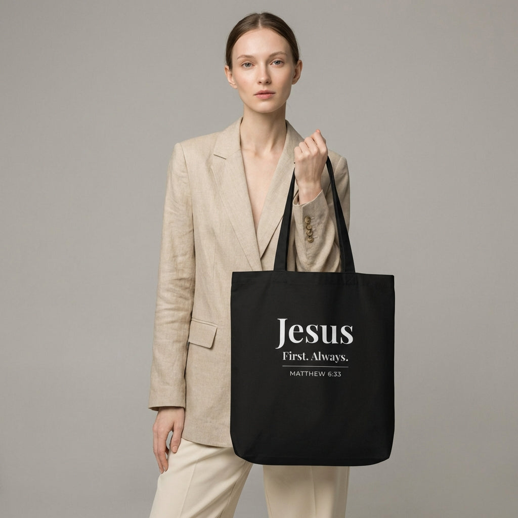 Model holding a black cotton tote bag printed with “Jesus First. Always.” and “Matthew 6:33” in white text.