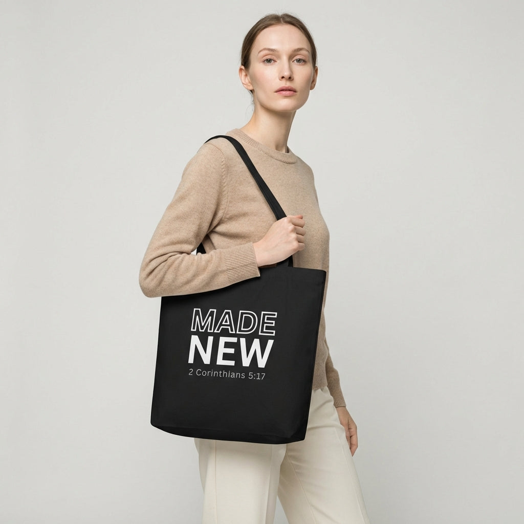 Model holding a black cotton tote bag printed with “MADE NEW” and “2 Corinthians 5:17” in white text.