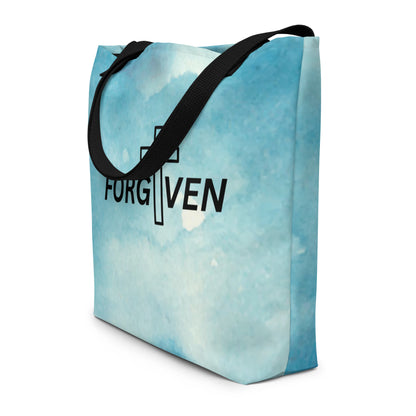 Angled view of light blue watercolor tote bag with black straps and “FORGIVEN” text with a cross detail.