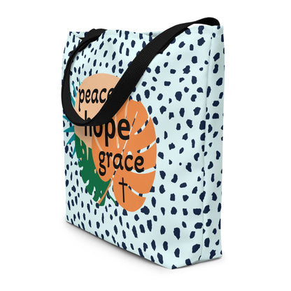 Angled view of light blue speckled tote bag with black straps and “peace hope grace” tropical leaf graphic with a small cross.