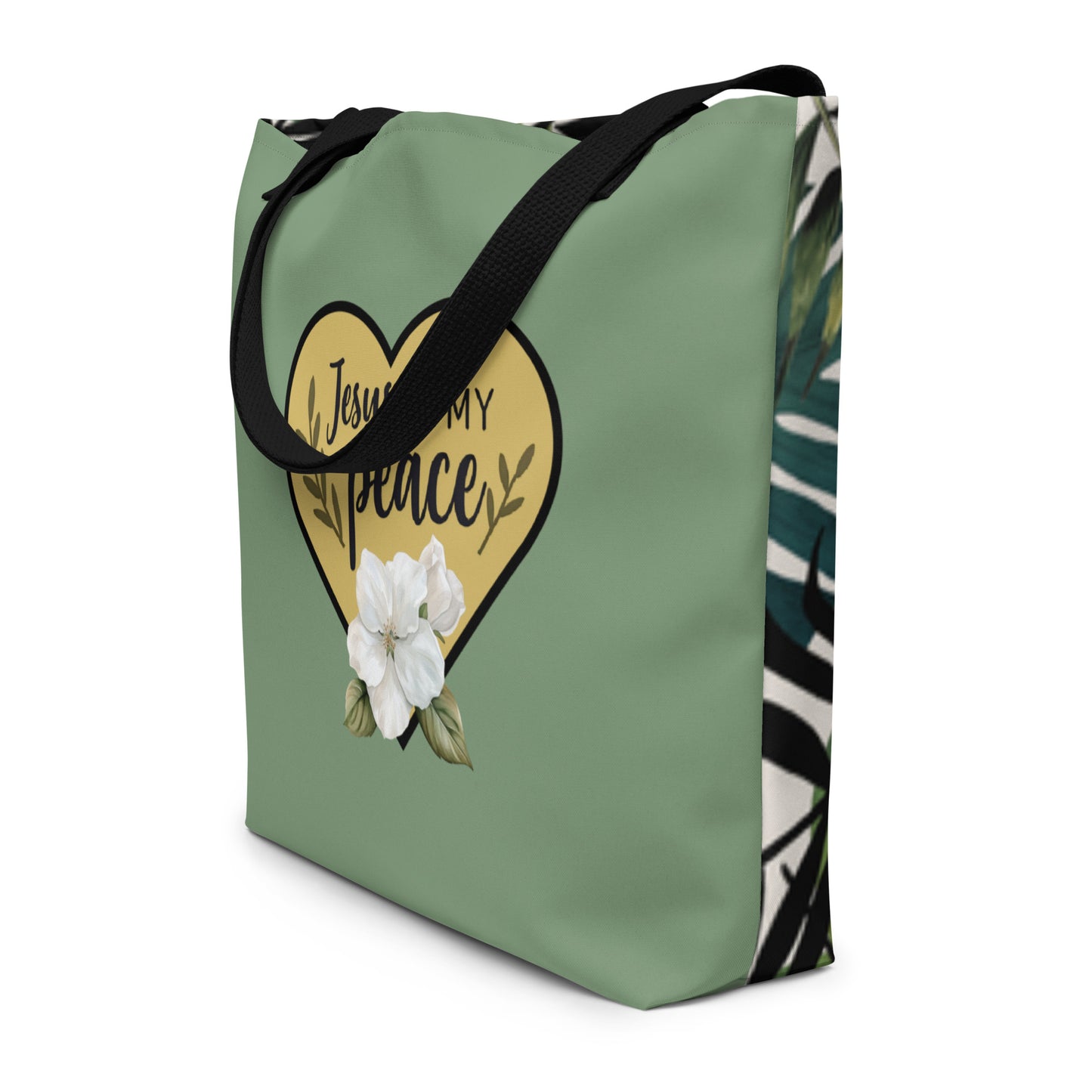 Angled view of the tote showing black straps and the wraparound design—sage green front with the gold heart “Jesus is my peace” graphic, and a tropical leaf pattern visible on the side/back.