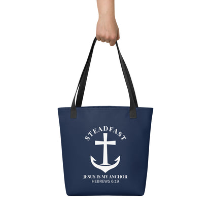 Tote bag held by hand with black straps, featuring a navy background and a white anchor cross graphic with the words “STEADFAST” and “JESUS IS MY ANCHOR” and the verse “HEBREWS 6:19.”