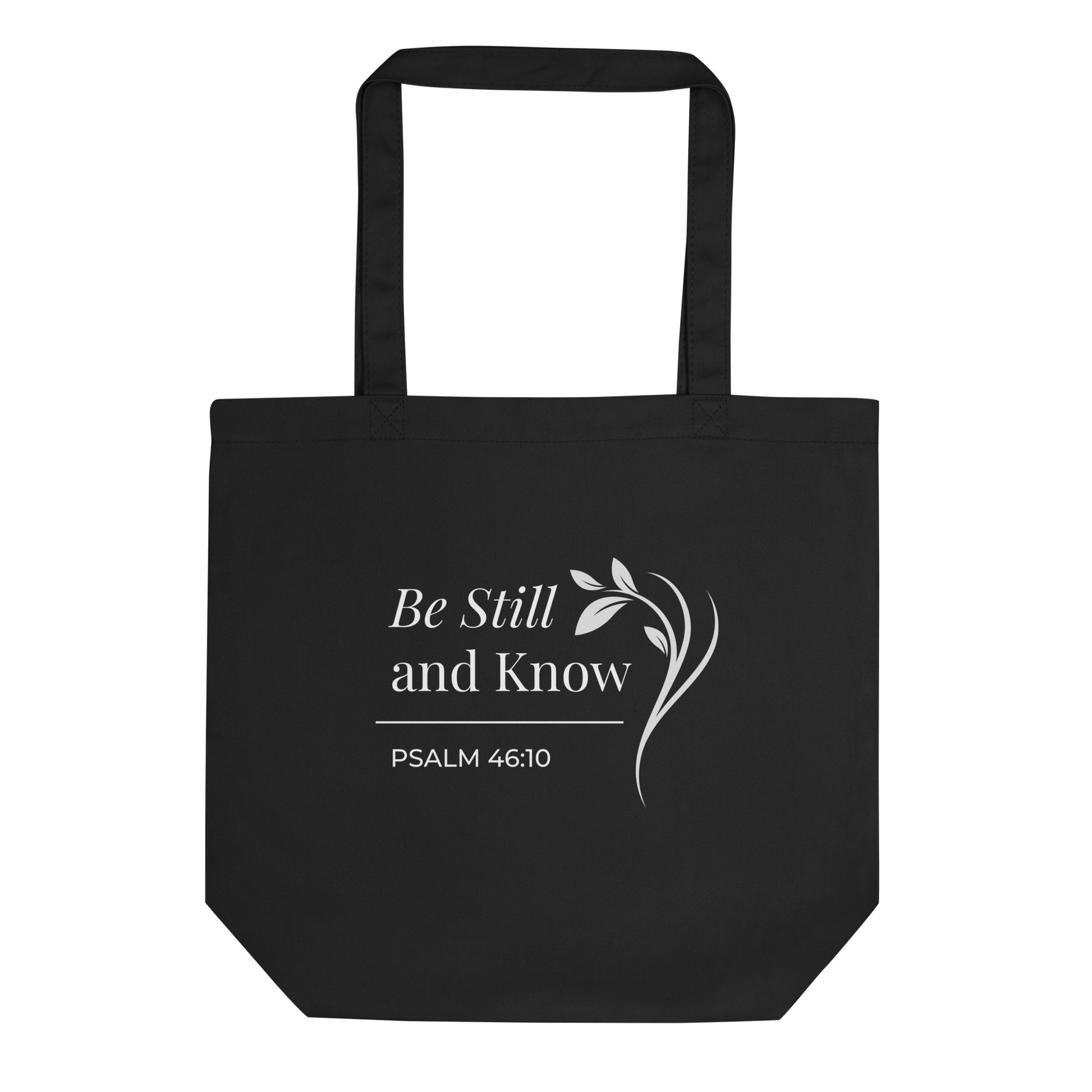 Front view of a black cotton Christian tote bag with matching black handles, featuring white “Be Still and Know” lettering, Psalm 46:10, and a graceful leafy branch design.