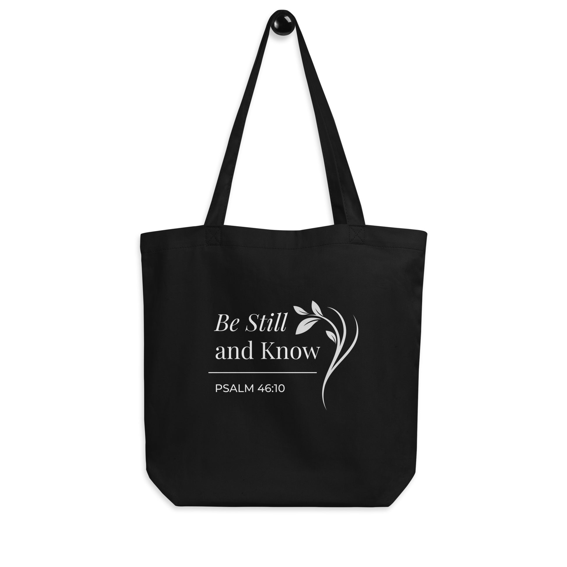 Black Christian cotton tote bag hanging on a hook, featuring matching black straps, Psalm 46:10 scripture text, and a simple curved leafy branch design.