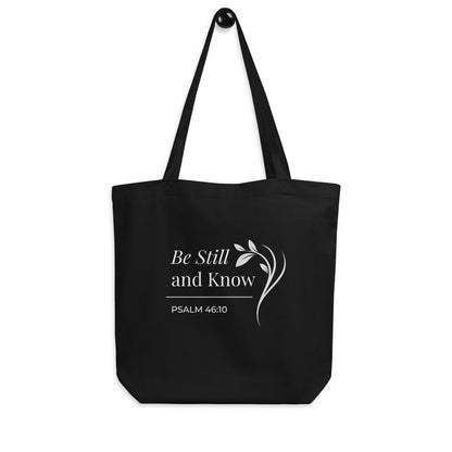 Black Christian cotton tote bag hanging on a hook, featuring matching black straps, Psalm 46:10 scripture text, and a simple curved leafy branch design.