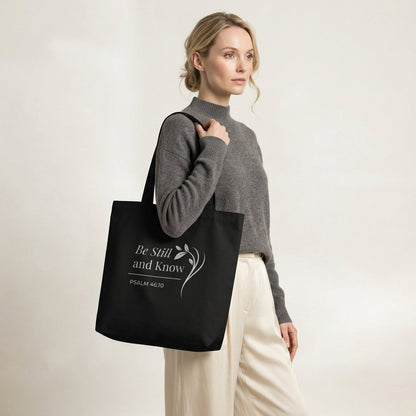Model carrying a black cotton Christian tote bag with matching black handles, white “Be Still and Know” lettering, and a delicate leafy branch illustration.