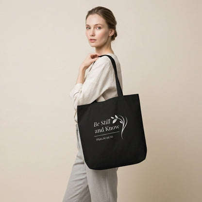 Model carrying a black cotton Christian tote bag with matching black handles, white “Be Still and Know” lettering, and a delicate leafy branch illustration.