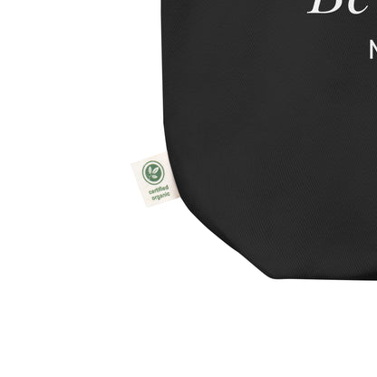 Close-up of a black cotton Christian tote bag showing the fabric texture, certified organic side tag, and part of the white “Be the Light” design.