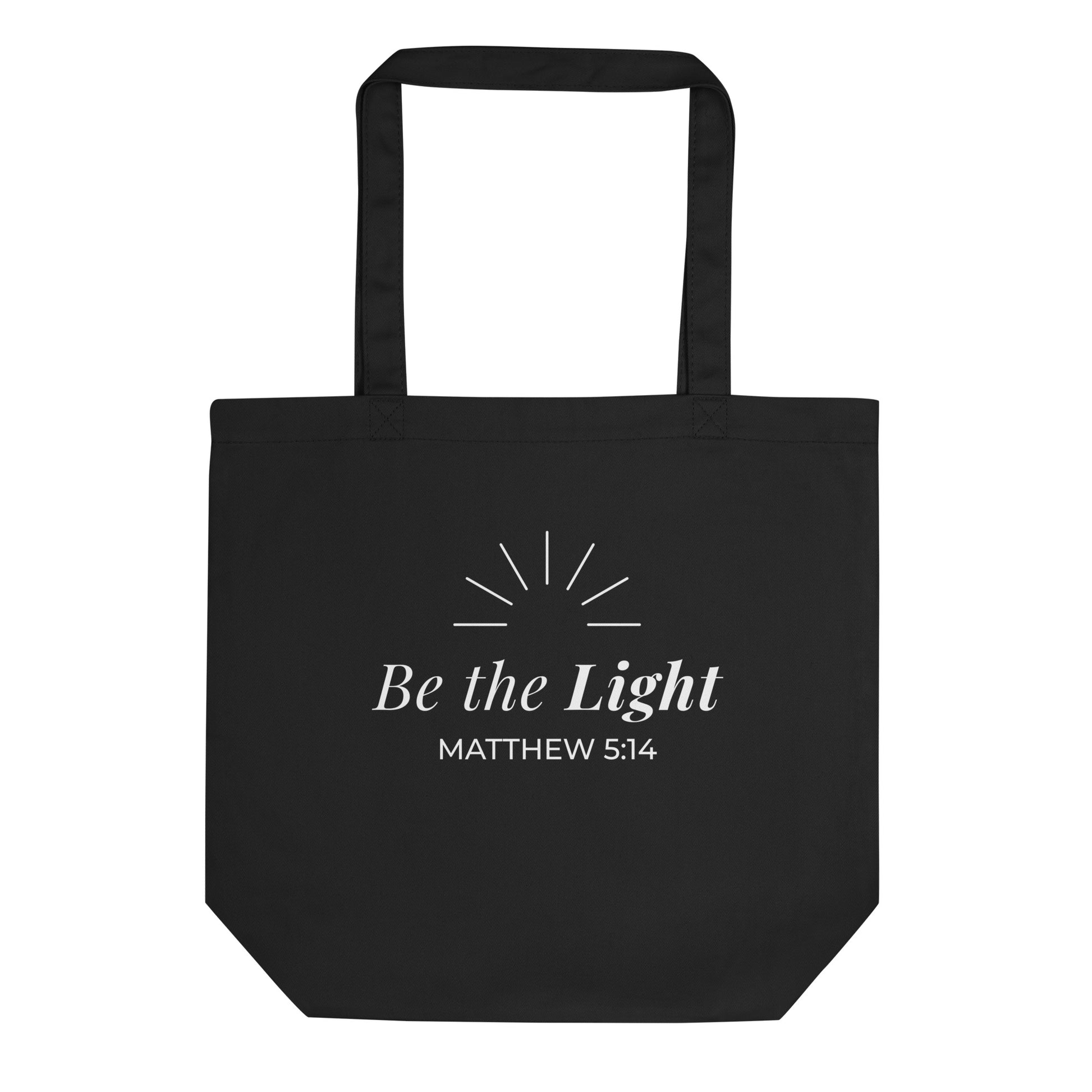 Front view of a black cotton Christian tote bag with matching black handles, featuring white “Be the Light” lettering, Matthew 5:14, and simple light ray artwork above the text.