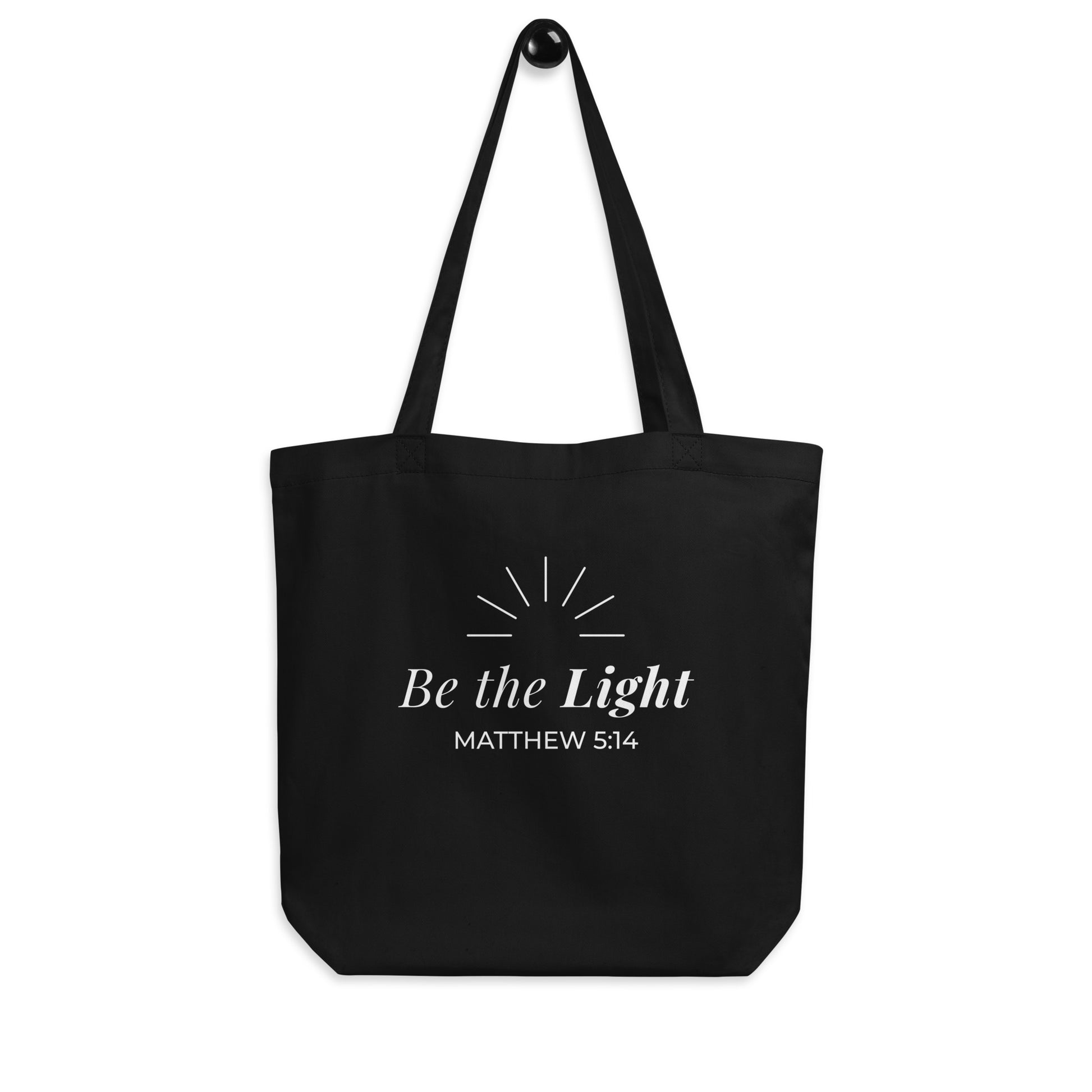 Black Christian cotton tote bag hanging on a hook, featuring matching black straps, white “Be the Light” text, and simple radiating light lines.
