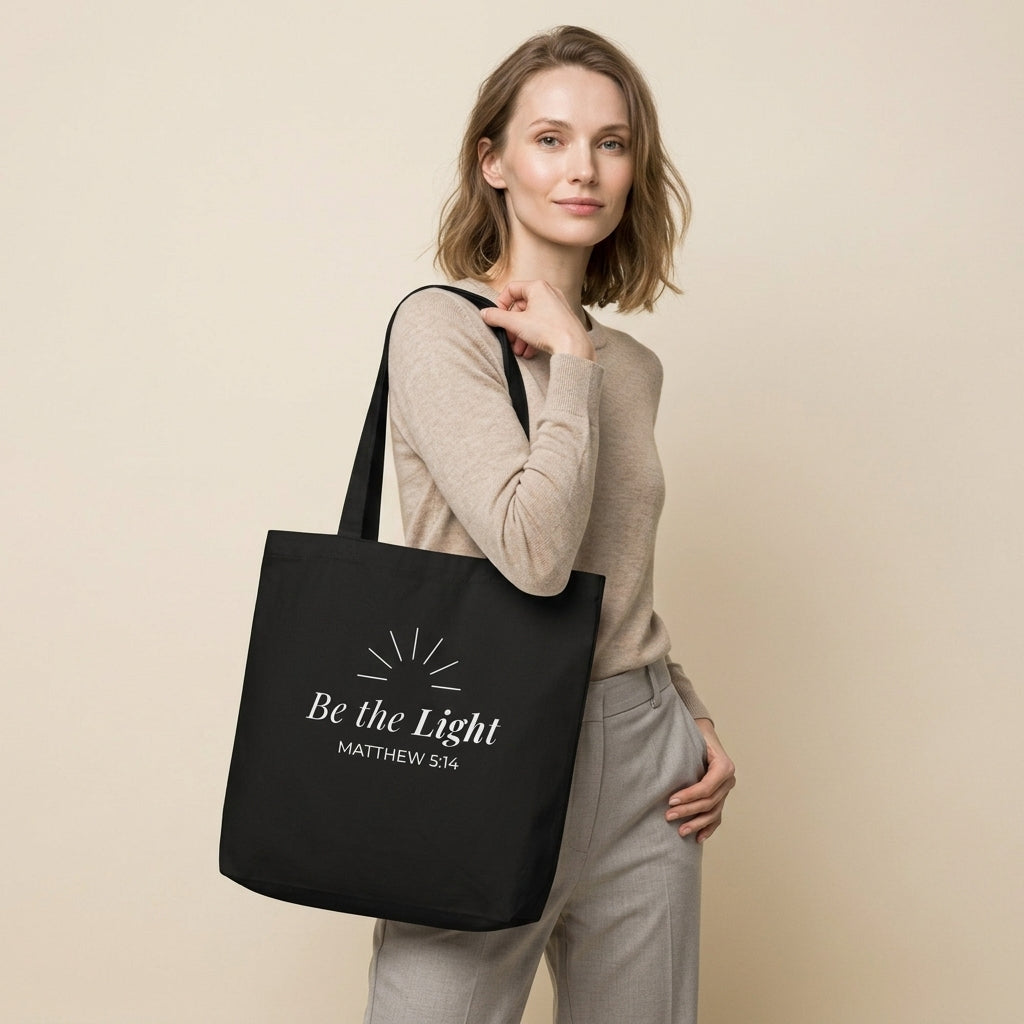 Model carrying a black cotton Christian tote bag with matching black handles and a minimalist “Be the Light” design inspired by Matthew 5:14.