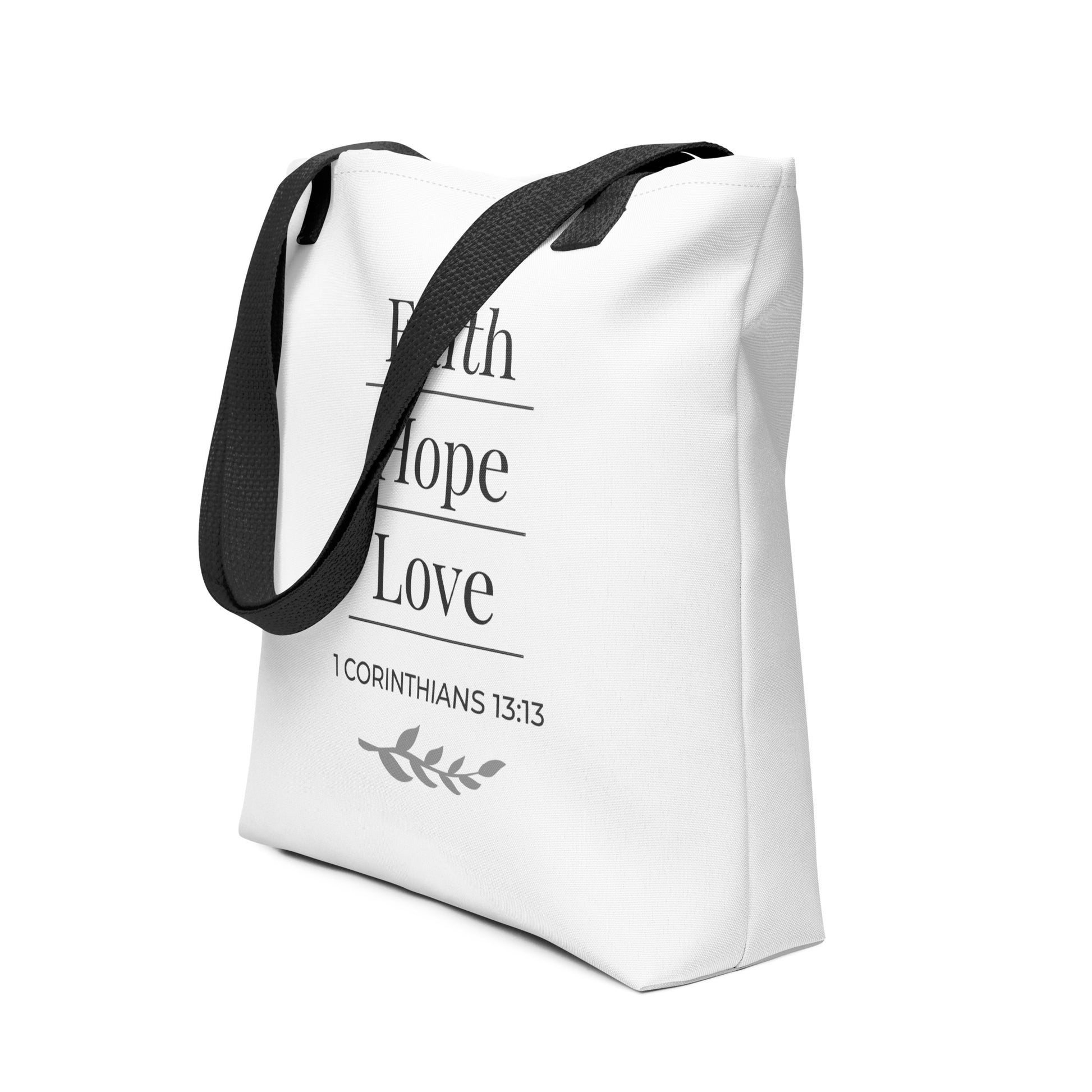 Angled view of a white tote bag with black handles featuring “Faith Hope Love,” “1 Corinthians 13:13,” and a leaf design in dark gray.