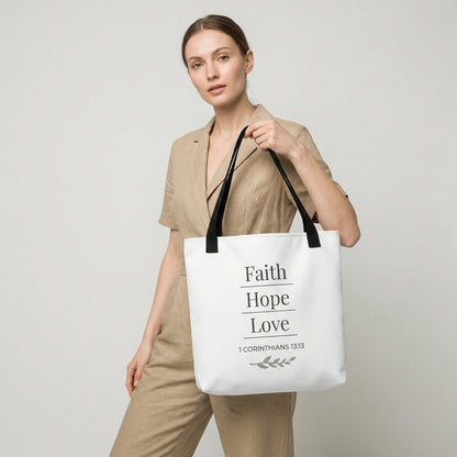 Lifestyle photo of a model holding a white tote bag with black handles featuring “Faith Hope Love,” “1 Corinthians 13:13,” and a leaf design in dark gray.