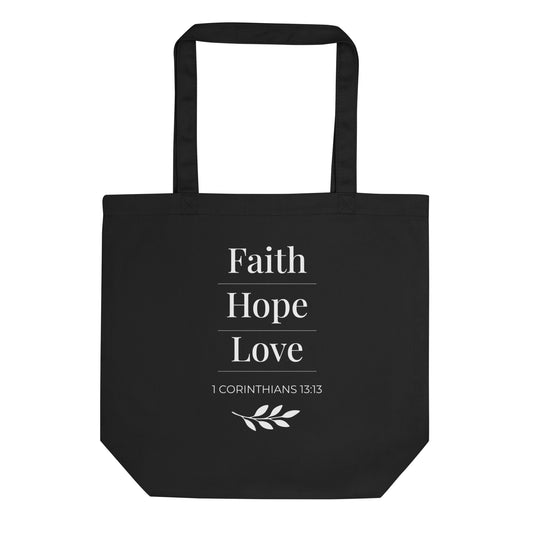 Front view of a black cotton Christian tote bag with matching black handles, featuring stacked white “Faith Hope Love” lettering, 1 Corinthians 13:13, underline accents, and a small leafy branch design.