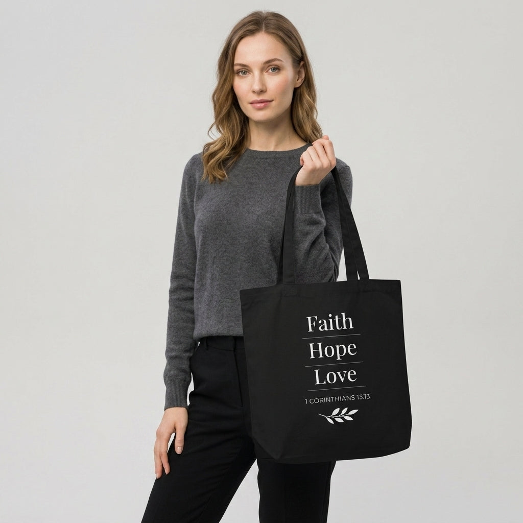 Model carrying a black cotton Christian tote bag with matching black handles and a “Faith Hope Love” 1 Corinthians 13:13 design with a small leafy branch illustration.
