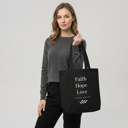 Model carrying a black cotton Christian tote bag with matching black handles and a “Faith Hope Love” 1 Corinthians 13:13 design with a small leafy branch illustration.