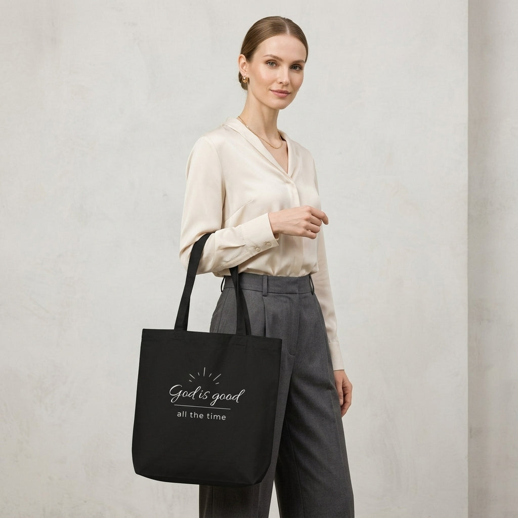 Model carrying a black cotton Christian tote bag with matching black handles and a minimalist “God is good all the time” faith design.