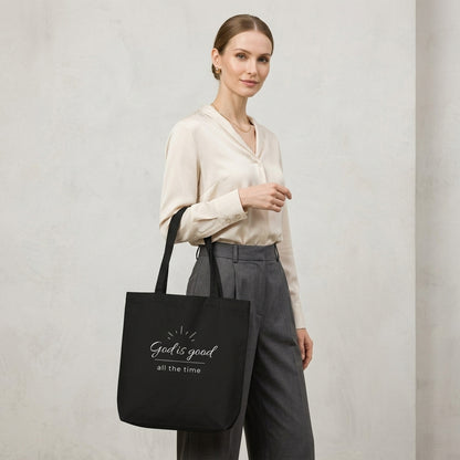 Model carrying a black cotton Christian tote bag with matching black handles and a minimalist “God is good all the time” faith design.