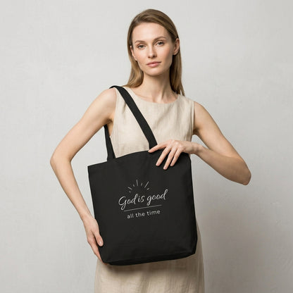 Model carrying a black cotton Christian tote bag with matching black handles and a minimalist “God is good all the time” faith design.