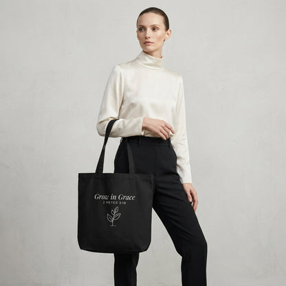 Model carrying a black cotton Christian tote bag with matching black handles, a “Grow in Grace” scripture design, and a small sprouting plant illustration.