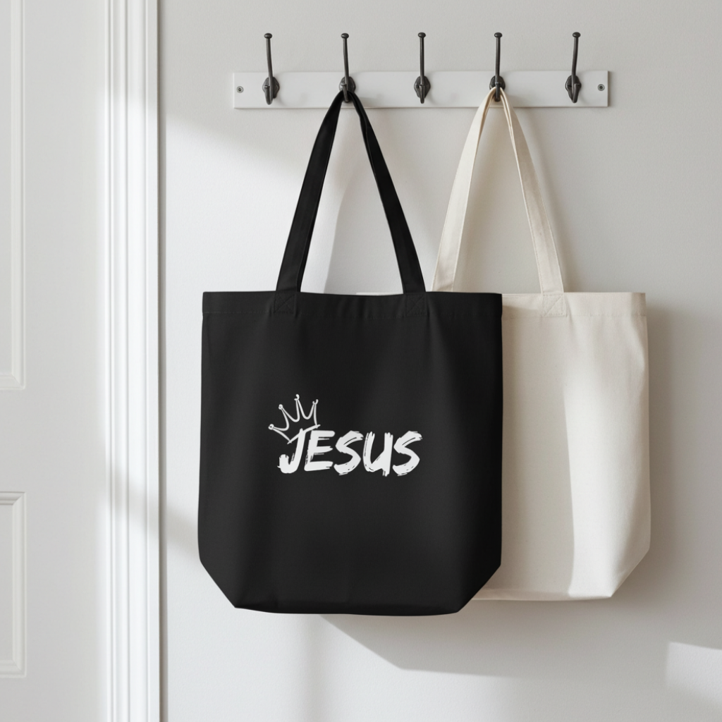 Black tote bag with 'JESUS' text and white tote bag hanging on a hook against a white wall.