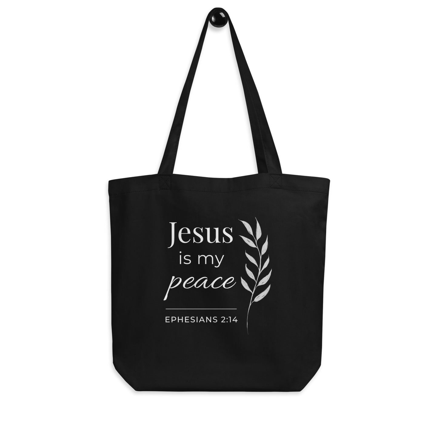 Black Christian cotton tote bag hanging on a hook, featuring matching black straps, white “Jesus is my peace” text, and a tall leafy branch design.
