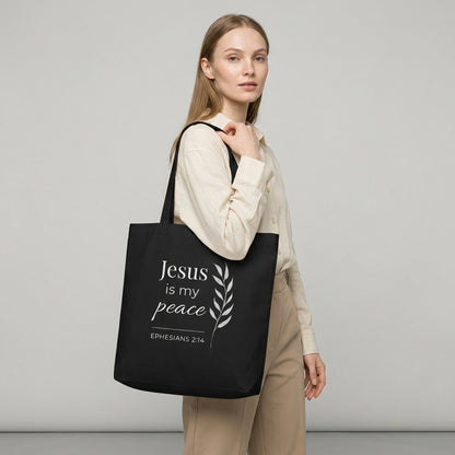 Model carrying a black cotton Christian tote bag with a minimalist “Jesus is my peace” design, Ephesians 2:14, and a graceful white leafy branch illustration.