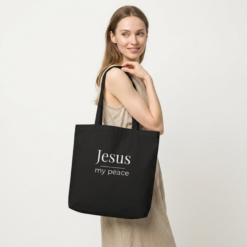 Model carrying a black cotton Christian tote bag with matching black handles and a minimalist “Jesus my peace” faith design.