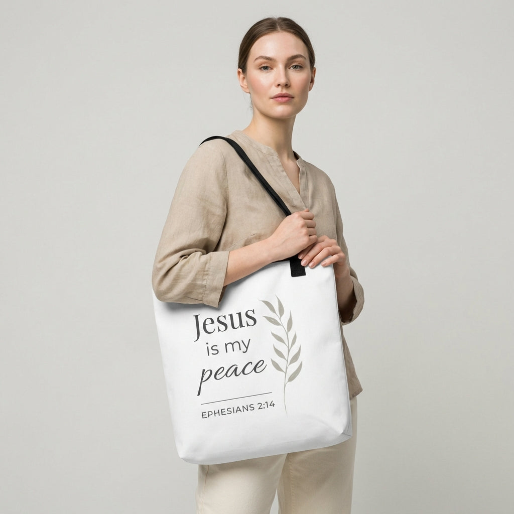 Lifestyle photo of a model holding a white tote bag with black handles featuring “Jesus is my peace,” “Ephesians 2:14,” and a leaf design in dark gray.