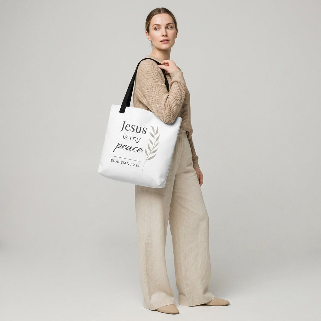 Lifestyle photo of a model holding a white tote bag with black handles featuring “Jesus is my peace,” “Ephesians 2:14,” and a leaf design in dark gray.