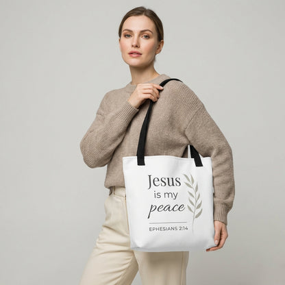 Lifestyle photo of a model holding a white tote bag with black handles featuring “Jesus is my peace,” “Ephesians 2:14,” and a leaf design in dark gray.