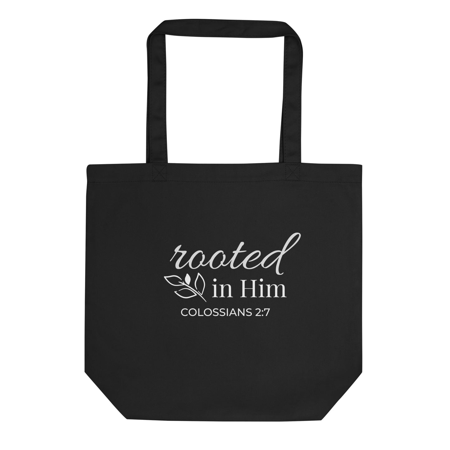 Front view of a black cotton Christian tote bag with matching black handles, featuring white “Rooted in Him” lettering, Colossians 2:7, and a small leafy branch accent.