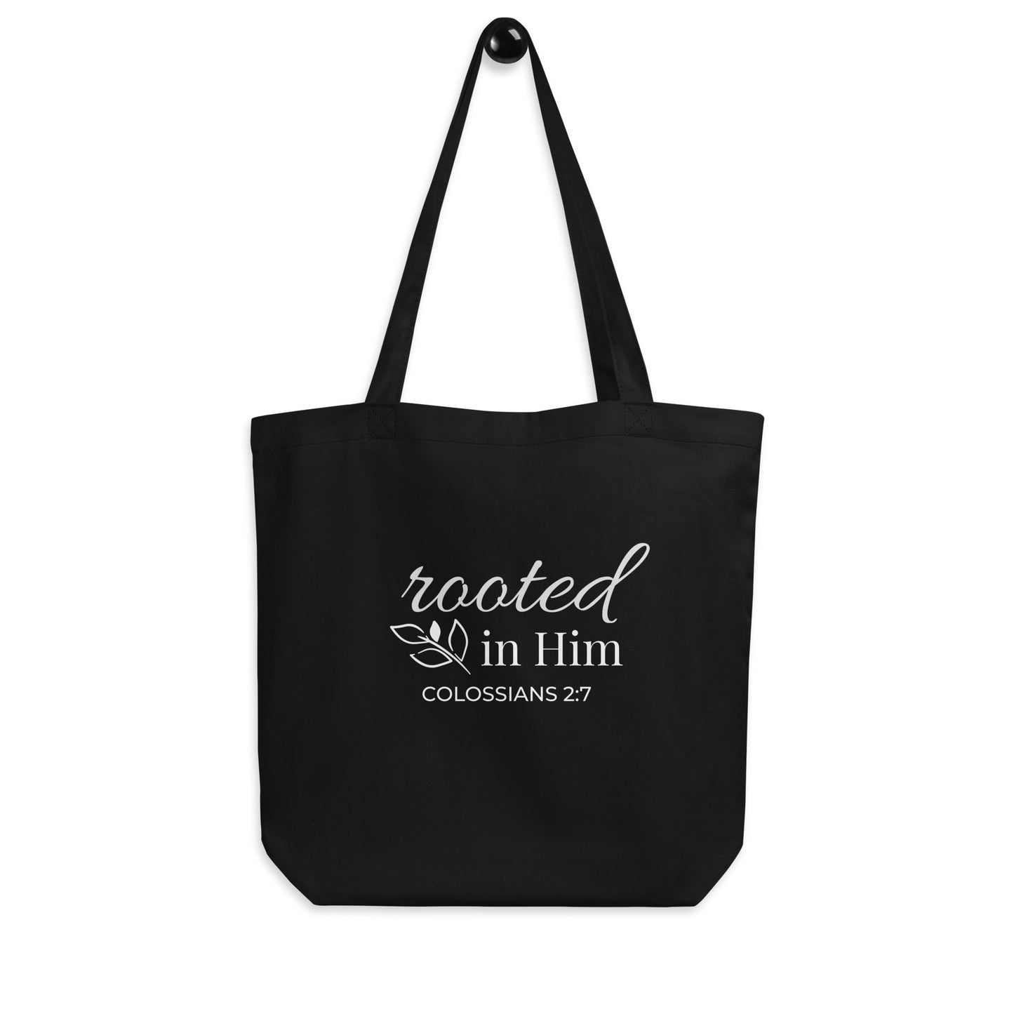 Black Christian cotton tote bag hanging on a hook, featuring matching black straps, white “Rooted in Him” text, and a small botanical leaf accent.