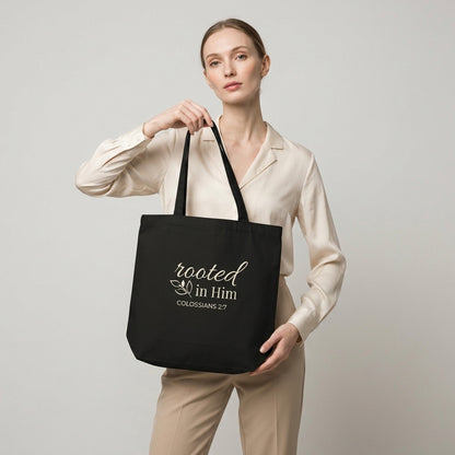 Model carrying a black cotton Christian tote bag with matching black handles, a “Rooted in Him” Colossians 2:7 design, and a simple leafy branch illustration.