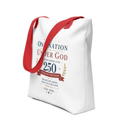 Angled view of a Christian Fourth of July tote bag featuring red handles, patriotic red, white, blue, and gold lettering, and “One Nation Under God.”