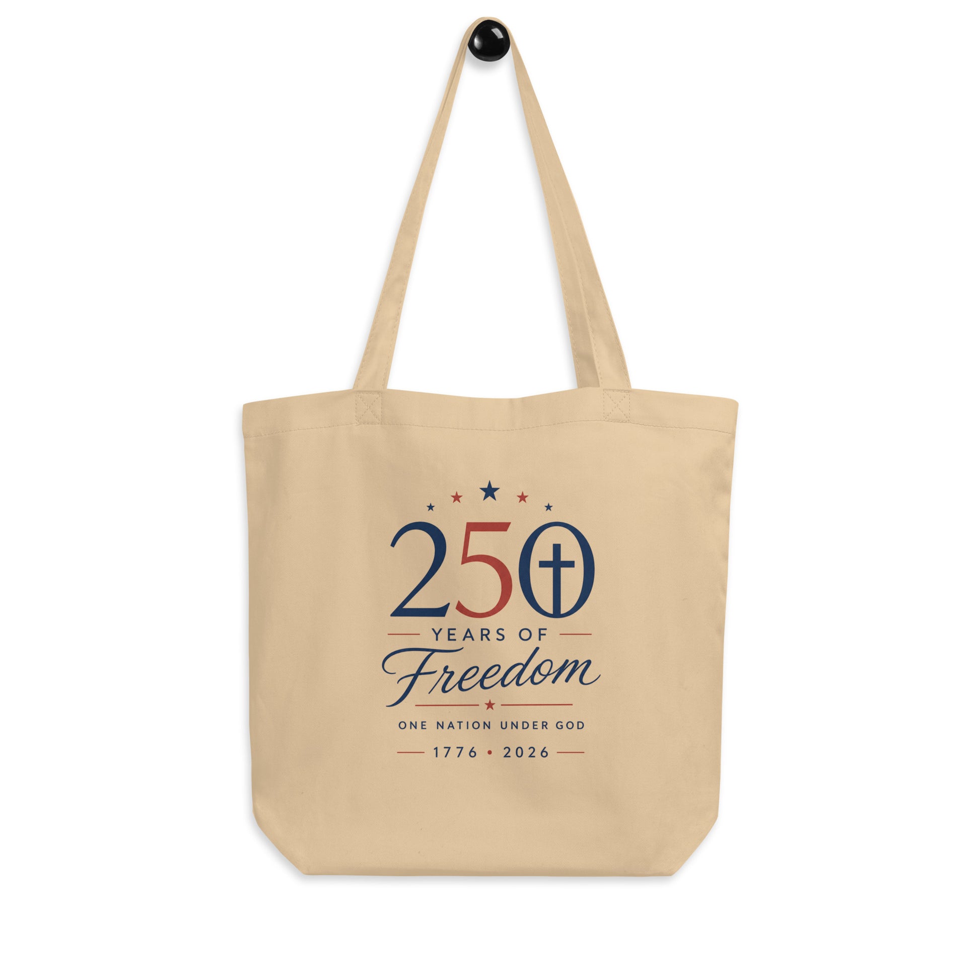 Christian patriotic cotton tote bag hanging on a hook, featuring a neutral beige color, red and blue artwork, and a cross design.