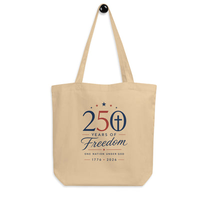 Christian patriotic cotton tote bag hanging on a hook, featuring a neutral beige color, red and blue artwork, and a cross design.