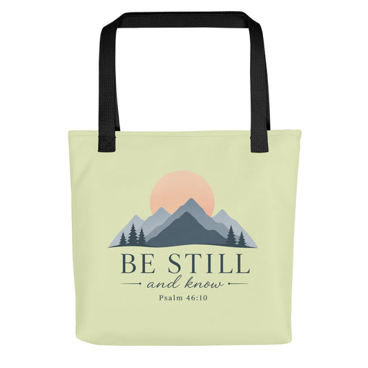 Front view of a light green Christian tote bag with black handles, mountain artwork, a sunrise, and the text “Be Still and Know Psalm 46:10.”
