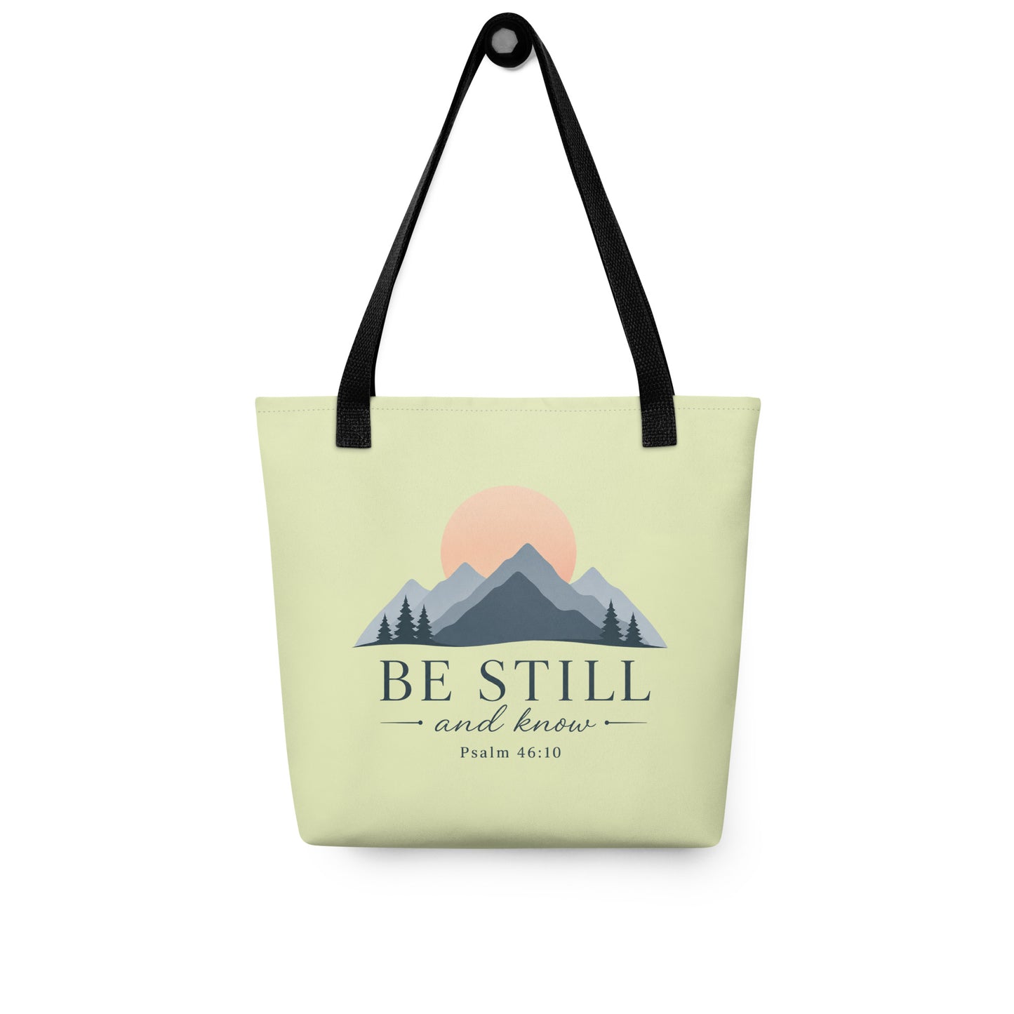 Christian tote bag hanging on a hook, featuring black straps, a soft green body, mountain artwork, and “Be Still and Know” lettering.