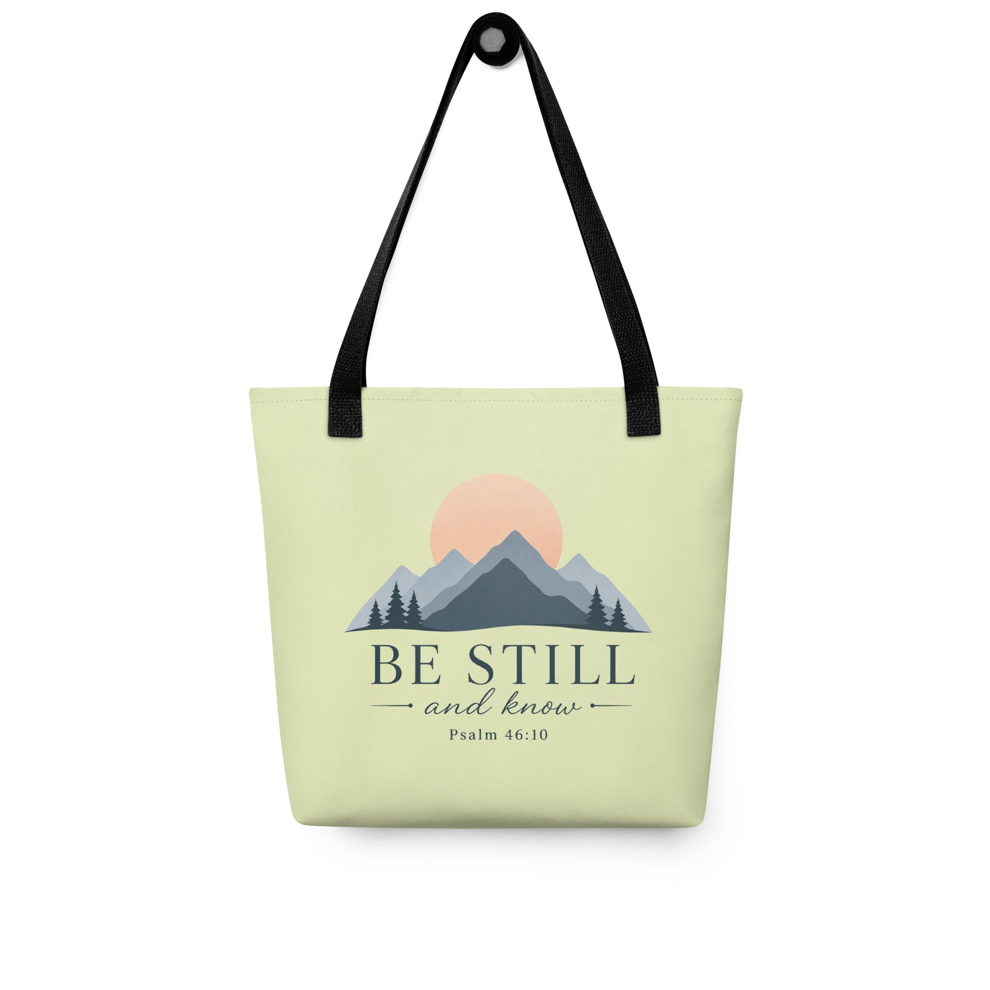 Christian tote bag hanging on a hook, featuring black straps, a soft green body, mountain artwork, and “Be Still and Know” lettering.