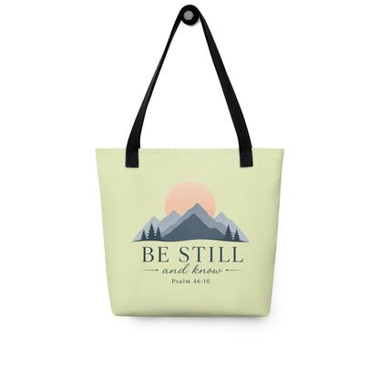 Christian tote bag hanging on a hook, featuring black straps, a soft green body, mountain artwork, and “Be Still and Know” lettering.