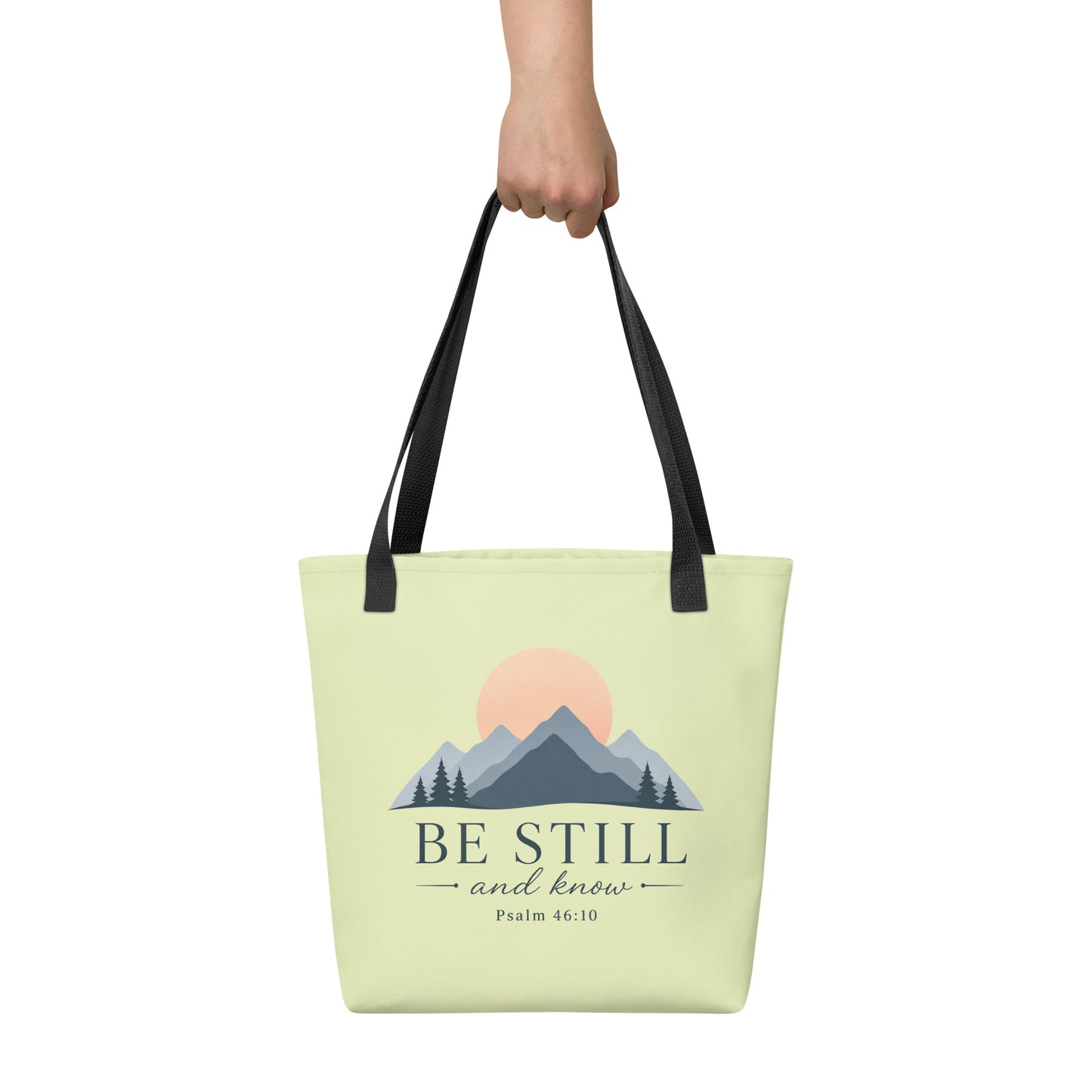 Person holding a light green Christian tote bag with black handles, a mountain sunrise design, and the Bible verse reference Psalm 46:10.