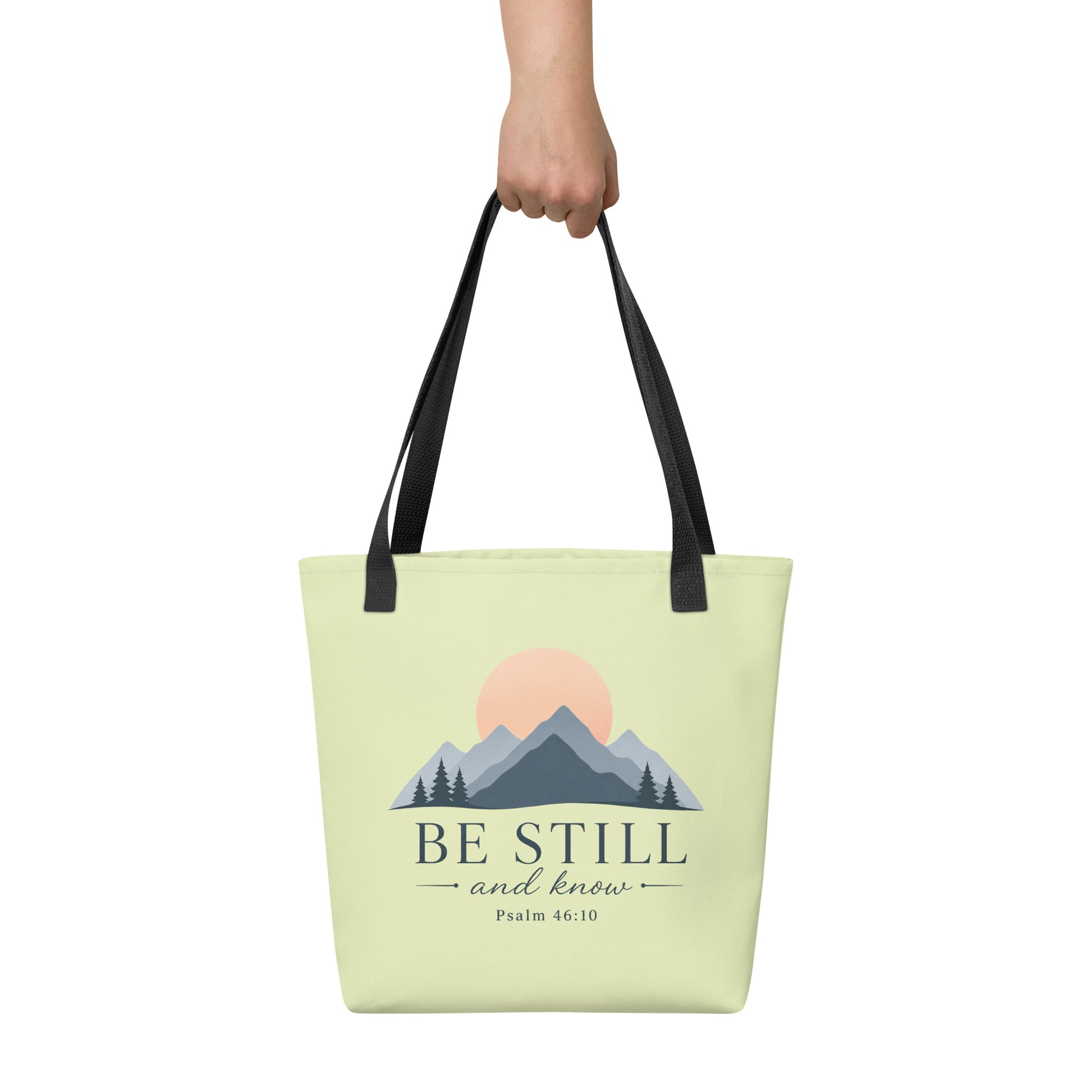 Person holding a light green Christian tote bag with black handles, a mountain sunrise design, and the Bible verse reference Psalm 46:10.