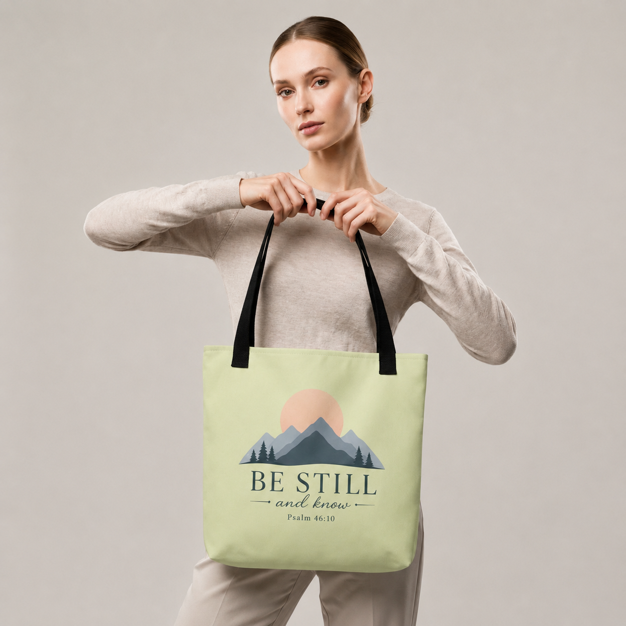 Model carrying a light green Christian tote bag with black handles and a peaceful mountain design featuring Psalm 46:10.