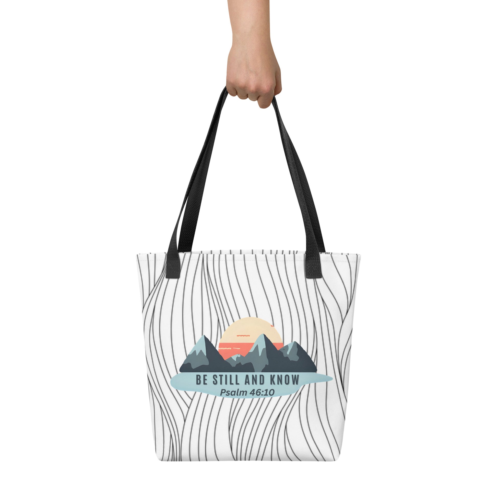 Tote bag held in hand with black handles featuring the mountain-and-sun artwork and “BE STILL AND KNOW — Psalm 46:10.”