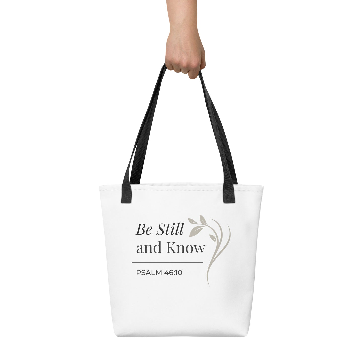 White tote bag held in hand with black handles featuring “Be still and know,” “Psalm 46:10,” and a floral line design in dark gray.