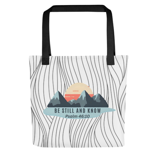 Front view of a patterned tote bag with black handles featuring a mountain-and-sun graphic and the text “BE STILL AND KNOW” with “Psalm 46:10.”