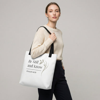 Lifestyle photo of a model holding a white tote bag with black handles featuring “Be still and know,” “Psalm 46:10,” and a floral line design in dark gray.